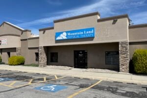 Exterior view of Mountain Land Physical Therapy clinic with a blue sign on a beige stucco building, stone pillars, and a blue handicap parking symbol in the foreground under a clear blue sky.
