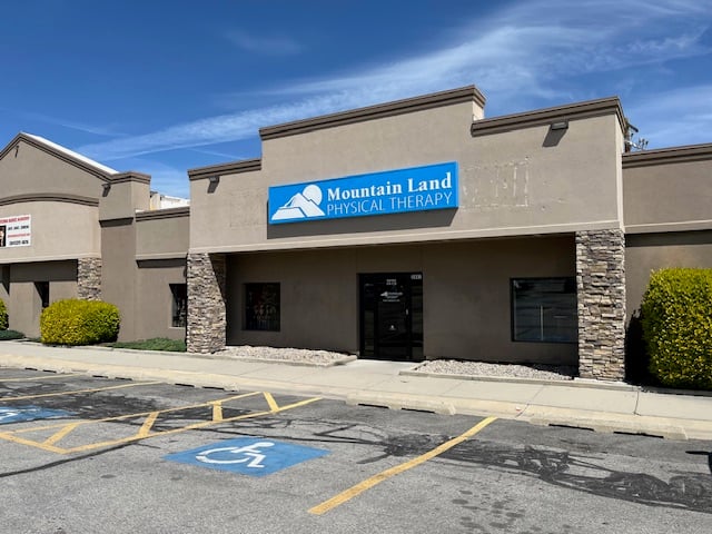 Exterior view of Mountain Land Physical Therapy clinic with a blue sign on a beige stucco building, stone pillars, and a blue handicap parking symbol in the foreground under a clear blue sky.