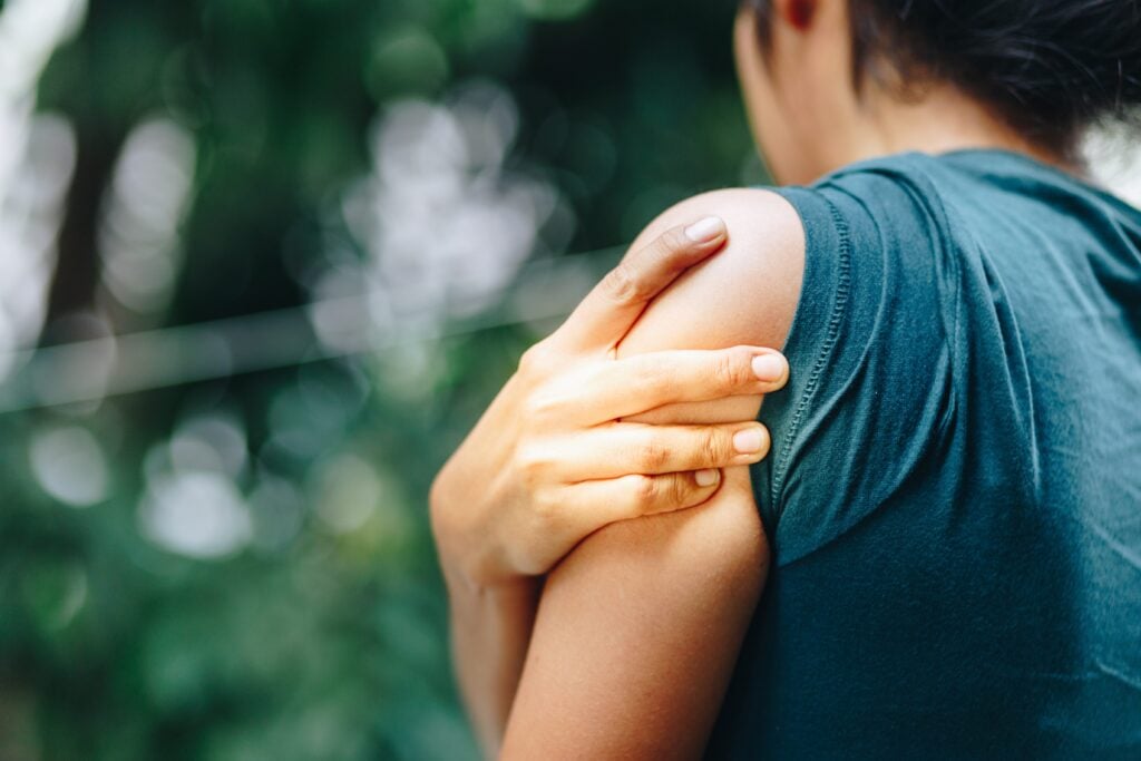 A close-up shot of a young woman experiencing shoulder pain. She is seen from behind, gripping her shoulder. She is wearing a green-grey shirt. She is outdoors.