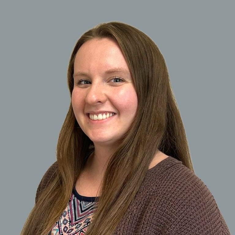 Smiling woman with long brown hair wearing a patterned top and brown cardigan against a gray background.