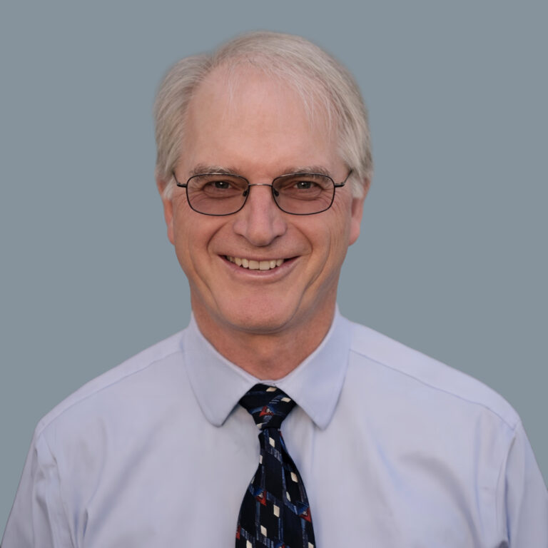 Smiling older man with gray hair and glasses, wearing a light blue dress shirt and striped tie against a blue-gray background.