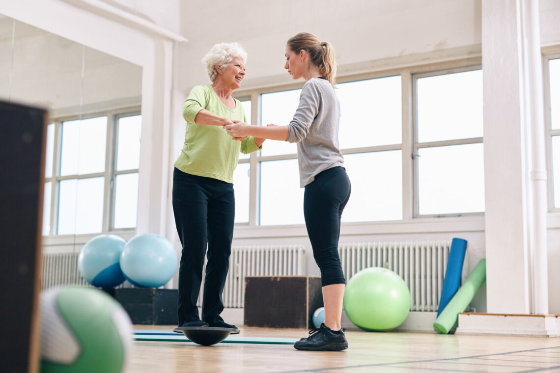 Female trainer helping senior woman in a gym exercising with a bosu balance training platform. Elder woman being assisted by gym instructor while workout session.