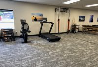Spacious fitness room with a treadmill, stationary bike, rowing machine, pull-up frame with orange bands, skeleton model, and wall photos.