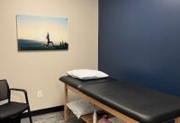 Treatment/therapy table in a small clinic room with a blue accent wall, pillow, and shelves of towels beneath the table.