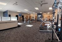 Rehabilitation gym with cardio machines (treadmill and elliptical), free weights, and therapy equipment scattered around a spacious, tiled room.
