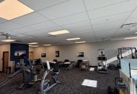 Large gym interior with multiple cardio machines (treadmills and bikes) in the foreground, free-weights and rack on the right, and wall photos along the back wall; sunlit through windows.