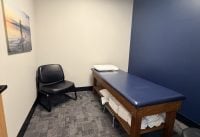 Small exam room with a blue padded treatment table along a dark blue wall, stacked towels beneath, and black chairs nearby with surf-themed wall art.