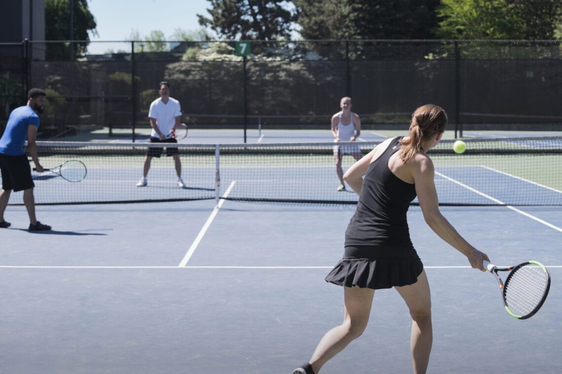 Four people playing tennis on outdoors court, a woman playing a forehand shot.