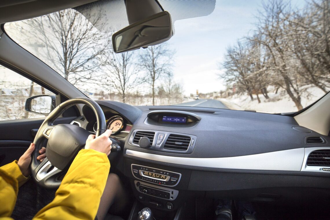Modern car interior with driver female hands on steering wheel, winter snowy landscape outside. Safe driving concept.