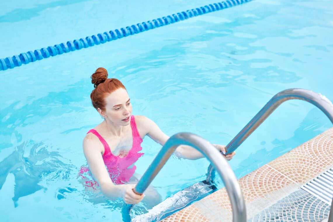tired cheerful girl standing in the swimming pool, doing exercises holding pool ladder,sportswoman, athlete relaxing in the swimming pool outdoors, close up side view photo